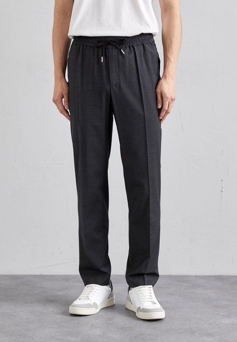 Homme portant un pantalon gris foncé à cordon et des baskets blanches, debout contre un mur uni gris clair.