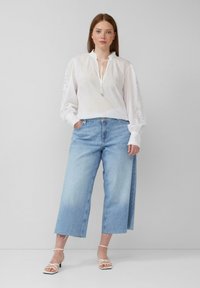 Chemisier blanc avec un col froncé et de longues manches bouffantes ; associé à un jean en denim bleu clair à jambes larges. Sandales à talons blancs.
