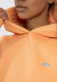 Perzikkleurige hoodie van zachte stof, met een ronde capuchon en een klein metallic logo aan de voorkant. Gladde textuur en eenvoudig ontwerp.