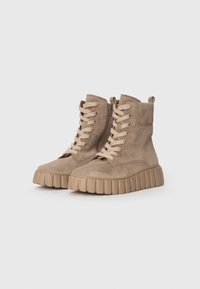 Une paire de bottines en daim beige avec de grosses semelles en caoutchouc ribdées, une fermeture éclair latérale et des lacets beiges sur un fond blanc uni.