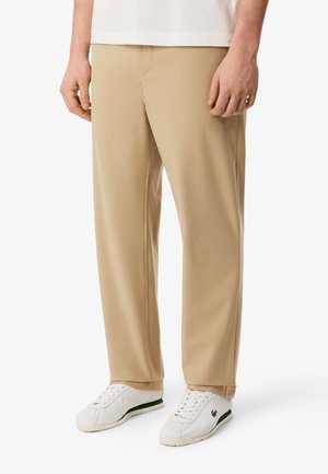 Homme portant un pantalon beige droit et des baskets blanches avec une bande verte, debout devant un fond blanc.