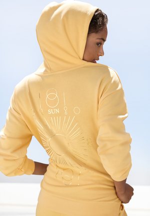 Yellow hooded sweatshirt with a soft texture, featuring metallic sun graphics and rays on the back. Front design is plain with a relaxed fit.