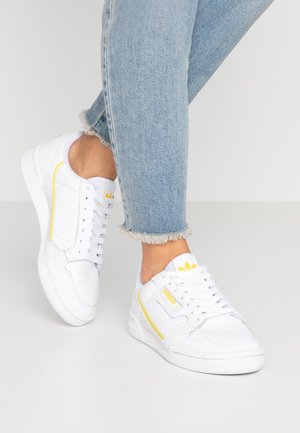 Person iført hvide Adidas sneakers med gule accenter og flossede lyseblå jeans, stående på en ensfarvet lys gulv.