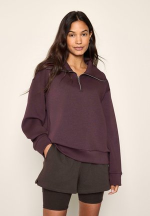 Donna con capelli lunghi e scuri che indossa una felpa viola scuro con zip e pantaloncini neri a strati con pantaloncini da ciclismo sotto.