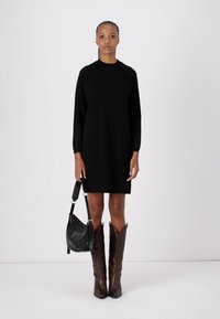 Melna kleita ar garām piedurknēm, brīvā piegriezumā, gluda tekstūra, apvienota ar melnu plecu somu un brūniem ādas kovboju zābakiem ar dekoratīvo dūrienu.