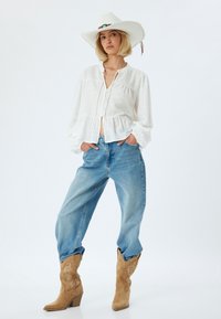 Blusa bianca con texture, con apertura frontale a bottoni e orlo a peplum, abbinata a jeans larghi di colore blu chiaro e stivali da cowboy beige con cuciture decorative.