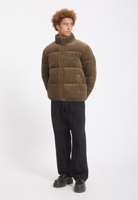 Jeune homme aux cheveux bouclés portant une veste matelassée marron, un pantalon noir et des chaussures noires, debout et regardant sur le côté.