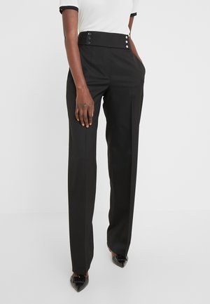 Vrouw die een zwarte broek met hoge taille en knoopdetails draagt, samen met een wit shirt met korte mouwen, staand met haar handen in de zakken.