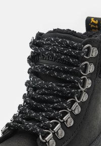 Cizme negre din suede, cu șireturi groase și texturate și gauri metalice. Tălpa interioară prezintă o etichetă cu marca "Dr. Martens" embosată.