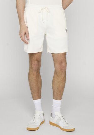 Man draagt witte shorts met trekkoord, witte enkelsokken en witte sneakers met gumzolen, staand tegen een effen lichtgrijze achtergrond.