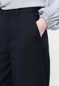 Marina-blå skräddarsydda byxor med slät tyg, rak skärning, sidofickor och en subtil lyster, sammansatta med en ljusgrå blus med veckade ärmslut.