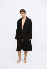 Melns peldmētelis no strukturētas auduma materiāla ar platu apkakli, garām piedurknēm, jostu viduklī un dizainu līdz ceļiem.