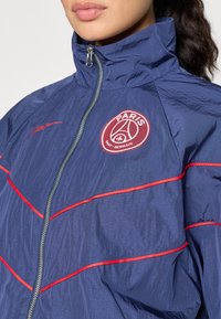 Námornícky vetrovka s červenými akcentmi a vysokým golierom. Má zips vpredu a vyšitý logotyp Paris Saint-Germain. Textúrovaná, hladká tkanina.