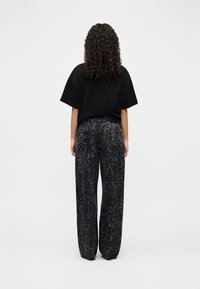 Γυναίκα με σγουρά μαλλιά, φορώντας μια μαύρη υπερμεγέθη μπλούζα και φαρδιά σκούρα γκρι παντελόνια με λεοπάρ σχέδιο, στέκεται με την πλάτη στραμμένη προς τα εμπρός σε απλό λευκό φόντο.