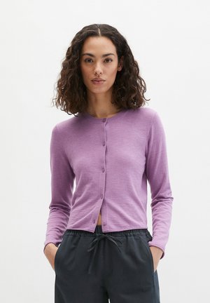 Frau mit lockigem Haar, die eine taillierte lavendelfarbene, geknöpfte Strickjacke und schwarze Jogginghosen mit Kordelzug trägt, steht mit den Händen in den Taschen.