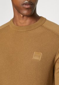 Brun stickad tröja med ribbat mönster, rund halsringning och en rektangulär läderlapp med texten "BOSS" på bröstet.