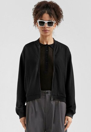 Zwarte mesh bomberjack met geribbelde boorden, ritssluiting, zijzakken, gedragen over een donker henley shirt, gecombineerd met witte zonnebril.