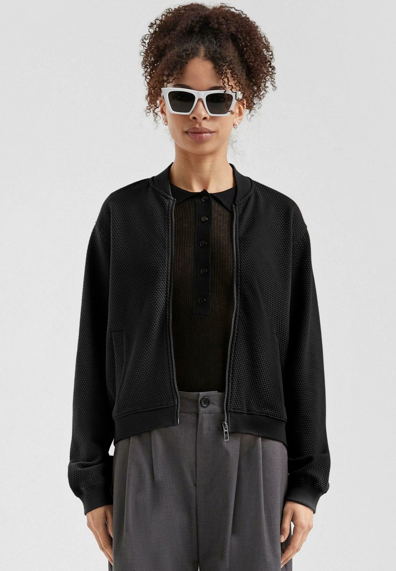 Zwarte mesh bomberjack met geribbelde boorden, ritssluiting, zijzakken, gedragen over een donker henley shirt, gecombineerd met witte zonnebril.