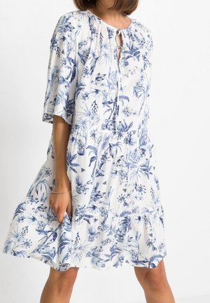 Femme portant une robe blanche à imprimé botanique bleu et manches courtes, tenant l'ourlet de la robe contre un fond uni.