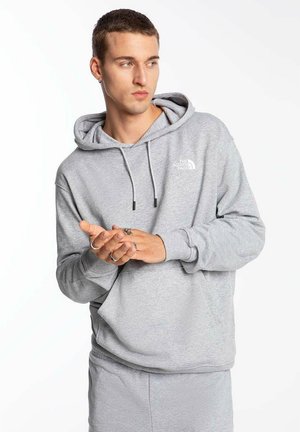 Light gray hoodie made of soft fabric, featuring a front pocket, drawstring hood, and small white logo on the left chest. Model showing hands.