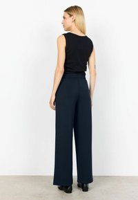 Woman with long blonde hair wearing a black sleeveless top, wide-leg navy pants, and black shoes, standing facing a white wall.