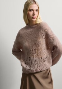 Pullover lavorato a maglia di un rosa chiaro con una vestibilità morbida, caratterizzato da un motivo in pizzo e maniche leggermente oversize, abbinato a una gonna liscia marrone.