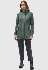 Grön fleecejacka med hög krage, dragkedjeförsedda sidofickor och kontrasterande sömnadsdetaljer, kombinerad med svarta jeans och stövlar.