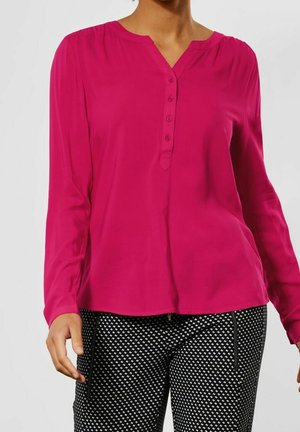 Femme portant un chemisier rose vif à manches longues avec des boutons et un pantalon noir avec un petit motif losange blanc.