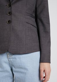 Blazer gris foncé avec cinq boutons noirs, coupe ajustée, texture subtile à fines rayures, et poches avant inclinées, associé à un denim clair.