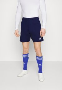 Pantalones cortos deportivos de color navy con cintura elástica, combinados con calcetas de fútbol a rayas azules y zapatillas blancas. El material parece ligero y transpirable.