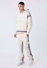 Giovane uomo con felpa crema e pantaloni della tuta con accenti grigi e sneakers bianche, in piedi davanti a uno sfondo chiaro e semplice.