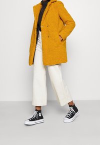 Cappotto teddy giallo a texture con cappuccio, due bottoni e tasche laterali, abbinato a pantaloni bianchi a gamba larga e sneakers alte nere.