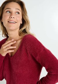 Pull à col en V, à manches longues, en rouge profond, fabriqué à partir d'un matériau doux et duveteux. Présente des poignets côtelés et une coupe décontractée.