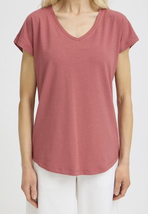 Žena odjevena u majicu kratkih rukava prljavo ružičaste boje s V-izrezom i bijele hlače, stoji nasuprot jednostavnoj svijetloj pozadini.