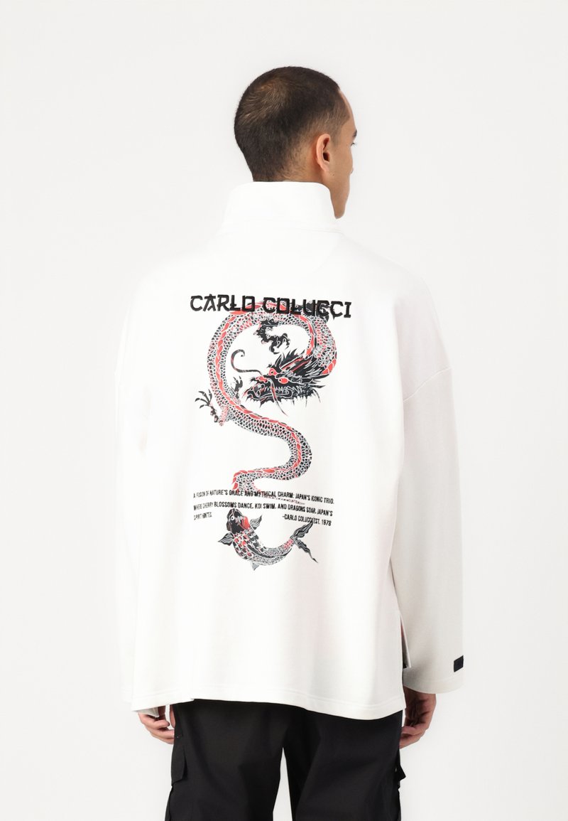 Biała, oversize koszula z grafiką smoka w czerwono-czarnych kolorach na plecach, z tekstem opisującym jej inspirację. Długie rękawy i boczne rozcięcia.