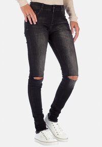 Jeans skinny neri con tagli strappati sulle ginocchia, vita alta e leggero sbiadimento. Abbinati a sneakers bianche, con design a lacci.