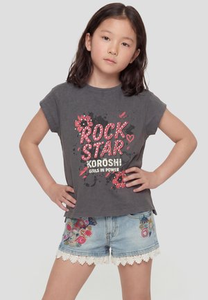 Niña joven con cabello oscuro de pie con las manos en las caderas, vistiendo una camiseta gris con la inscripción "Rock Star" y shorts de mezclilla bordados con borde de encaje.