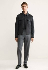 Chaqueta gris de mezcla de lana con cierre de botones, dos bolsillos en el pecho y cuello con solapa, combinada con pantalones grises a medida y zapatos negros.