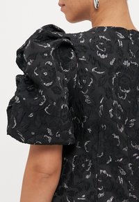 Čierne šaty so štruktúrovanými kvetinovými vzormi a metalickými akcentmi. Rukávy vo forme pufov a zips na chrbte obohacujú dizajn.