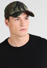 Tarnkappen-Basecap aus Stoff; mit einem schwarzen Logo auf der Vorderseite, gebogenem Schirm und leichter Textur, getragen mit einem schwarzen T-Shirt.