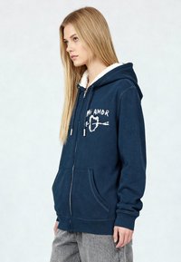 Sweat à capuche zippé bleu marine avec un col doublé en polaire blanc, comportant le texte "Mi Amore" et un design en forme de cœur. Comprend des poches à l'avant et des poignets côtelés.