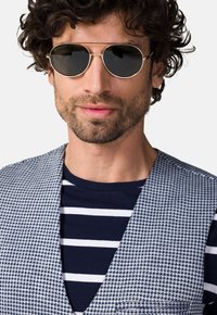 Męskie okulary przeciwsłoneczne z okrągłymi ciemnymi soczewkami i cienkimi złotymi oprawkami. Model ma na sobie niebieską koszulę w paski oraz teksturowaną szaro-białą kamizelkę.