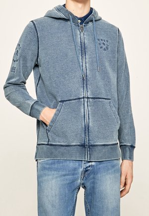Man draagt een vervaagde blauwe hoodie met ritssluiting, trekkoorden, voorzakken en lichtblauwe spijkerbroek, staat met één hand in de zak.