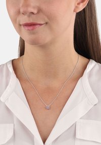 Kvinna som bär ett silverhalsband med en liten blomformad lila ädelsten som hänge, klädd i en vit blus.