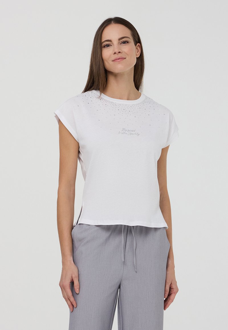 T-shirt bianco a maniche corte con scollatura rotonda, caratterizzato da un motivo di strass sparsi nella parte superiore e fessure laterali. Testo decorativo sotto il design.