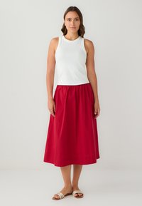 Top sans manches blanc associé à une jupe midi rouge. La jupe présente un design froncé et tombe gracieusement jusqu'aux chevilles, accompagnée de sandales légères.