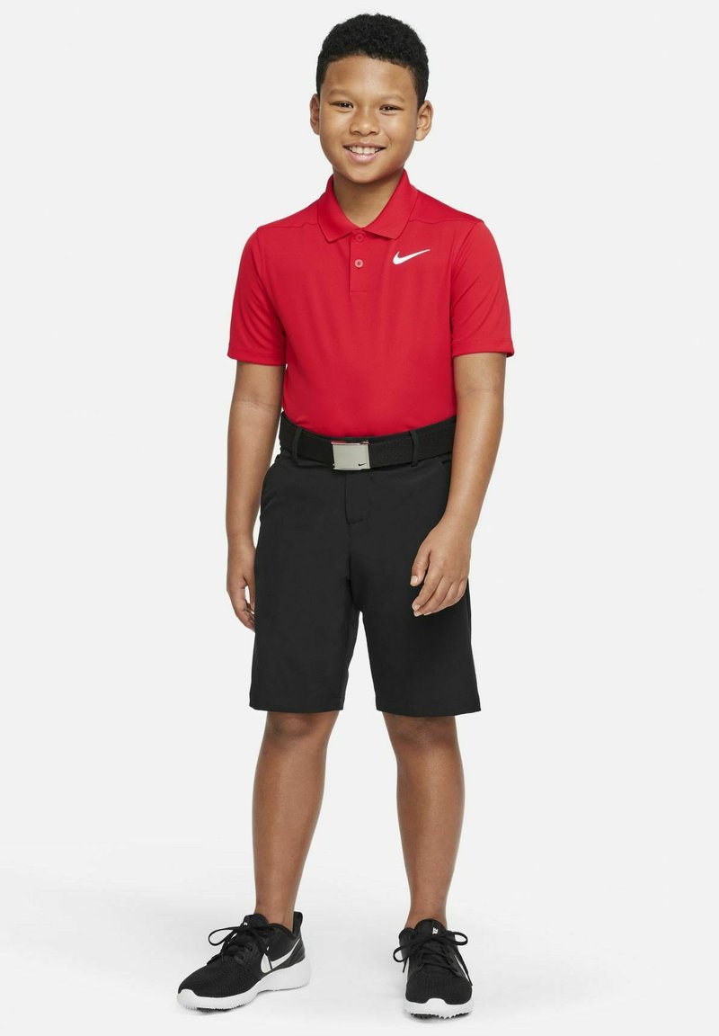 Czerwona koszulka polo z kołnierzykiem i logo Nike, czarne szorty, czarny pasek ze srebrną klamrą i czarne buty sportowe. Pozująca postawa.