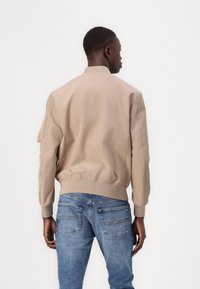 Beige bomberjakke med en glatt tekstur, elastiske mansjetter og hem, med sidelommer og et dempet design, båret over blå jeans.