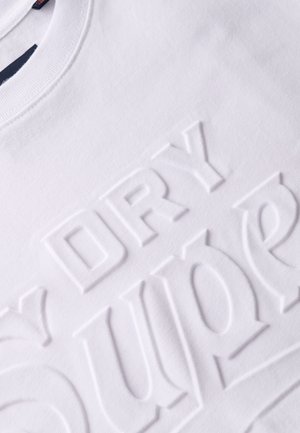 T-shirt en coton blanc avec des lettres en relief portant les mots "DRY" et "SUPER" dans une police moderne et audacieuse. Texture lisse, encolure ronde.