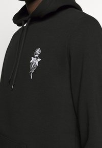 Hoodie noir avec main de squelette blanche brodée tenant une rose sur la poitrine, porté par une personne visible du cou au milieu du torse.